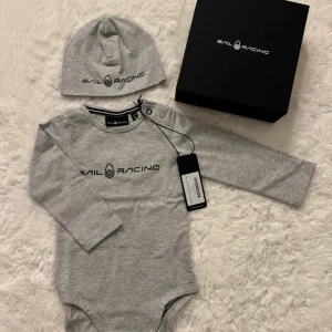 Sail Racing babyset grå 6-12 mån HELT NY i presentbok  - Sail Racing babyset, body och mössa i grått.   Setet består av en långärmad body med tryckknappar mellan benen och en matchande mössa med loggan.    Storlek 6–12 månader  100% bomull    🌟 Helt ny med tag kvar! 🌟   Levereras i original presentförpackning    perfekt som present! 🤍   Finns i fler färger och storlekar. Kolla mina andra annonser eller skriv medelanden.
