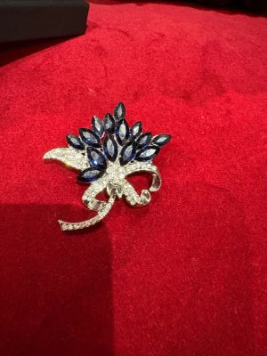 Blå glittrig brosch med strass - Säljer en riktigt snygg brosch i form av en blomma med mörkblå stenar och massor av glittrande strass. Broschen har en elegant rosettformad detalj och är perfekt för att piffa upp vilken outfit som helst. Fästet på baksidan gör den enkel att sätta på väst eller jacka.