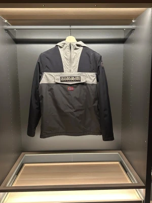 Napapijri anorak vindjacka - Snygg vindjacka från Napapijri i svart och grått med huva och halv dragkedja. Jackan har en stor logga på bröstet och norsk flagga, samt praktisk ficka framtill. Perfekt för dig som gillar streetwear och vill ha något som sticker ut. Hör av er vid frågor eller funderingar.