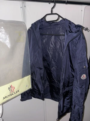 Mörkblå vindjacka från Moncler - Säljer en mörkblå vindjacka från Moncler med huva och dragkedja framtill. Jackan har Moncler-logga på ärmen och snygga detaljer som snörning vid huvan och fickor med knappar. Materialet är lätt och syntetiskt, perfekt för blåsiga dagar. Med skann.