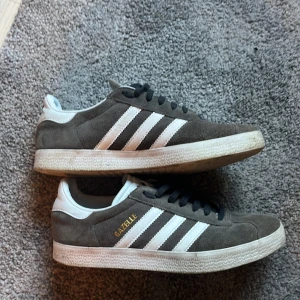 Adidas Gazelle grå mockasneakers - Säljer ett par klassiska Adidas Gazelle sneakers i grå mocka med vita detaljer och svarta snören. Skorna har inte används mycket! Skorna är 39 1/3 och passar både killar och tjejer! Säljer pga att de inte kommer till användning och för att de är för små skorna är i väldigt bra skick är lite smutsiga men kan tvätta innan jag fraktar! Skriv om ni undrar nåt! MVH Arvid 