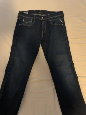 Replay jeans  - Snygga Replay Anbass jeans i mörkblå tvätt med kontrastsömmar och coola detaljer på bakfickorna. Slim fit-modell med klassisk femficksdesign och knappgylf. Tillverkade i jeansmaterial med återvunna fibrer för extra hållbarhet.