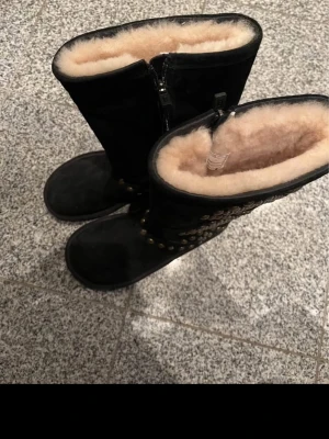 Svarta UGG boots med nitar och mönster - Säljer ett par svarta UGG boots i mocka med mjukt beige foder och platt sula. Snygga detaljer med nitar och diamantmönster på sidorna. Dragkedja på insidan för enkel påtagning. Perfekta för kalla dagar och riktigt sköna att ha på sig.