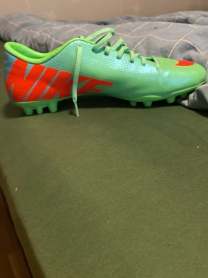 Nike Mercurial fotbollsskor mintgrön - Nike Mercurial fotbollsskor i mintgrön färg med starkt orange swoosh och logga. Skorna har snörning och platt sula med dobbar för gräsplan. Tillverkade i syntetmaterial med en sportig och modern look. Perfekt för dig som vill sticka ut på planen.