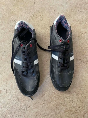 Svarta sneakers från Tommy Hilfiger - Snygga svarta sneakers från Tommy Hilfiger med vita och marinblå detaljer samt röd accent vid snörningen. Skorna har en platt sula, rund tå och snörning. Insidan har ett rutigt foder i blått, rött och vitt. Materialet är skinn och syntet.