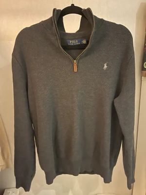 Grå half zip tröja från Polo Ralph Lauren - Snygg mörkgrå långärmad tröja från Polo Ralph Lauren med half zip och brunt läderdetalj på dragkedjan. Tröjan har broderad logga på bröstet och är tillverkad i bomull med stickad struktur. Perfekt för dig som gillar klassisk och stilren design.