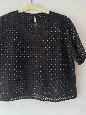 Svart blus  från Zara Woman - Säljer en svart blus från Zara Woman i storlek M. Blusen har korta ärmar och är tillverkad i ett genombrutet tyg med små  zirkon över hela ytan. Rund halsringning och knapp i nacken. Perfekt för dig som gillar unika detaljer och stilren design. 2st zirkon hänger, måste sys, se bilder, trådar förekommer