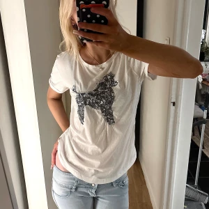 Vit t-shirt med paljettmotiv från Holly & Whyte - Vit t-shirt från Holly & Whyte med ett stort paljettmotiv av en hund på bröstet. Modellen är klassisk med rund hals och korta ärmar. Materialet är mjuk bomull som känns skönt mot huden. Perfekt för dig som gillar lite extra bling i vardagen.