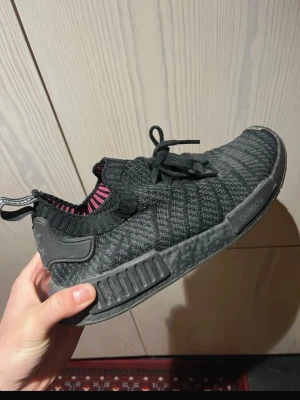 Svarta Adidas NMD sneakers - Säljer ett par svarta Adidas NMD sneakers med stickad ovandel och diskret randigt mönster. Skorna har svarta snören, platt sula och detaljer i gummi. Insidan har en touch av rosa. Perfekta för dig som gillar streetstyle och bekväma skor.