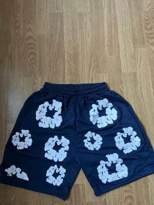 Blå shorts med blommigt tryck Denim Tears - Säljer ett par mörkblå shorts från Denim Tears i storlek M. De har elastisk midja med snörning och är dekorerade med stora vita blommönster över hela plagget. Materialet känns mjukt och lätt, perfekt för varma dagar.