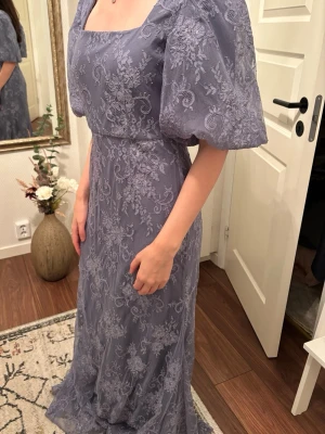 By Malina - spetsklänning - Elegant och romantisk lång klänning från By Malina i en mjuk blå/grå nyans med vackert genomgående spetsmönster. Klänningen har en stilren fyrkantig ringning och korta puffärmar. Smickrande midjesöm och ett följsamt fall som sitter fint och känns bekvämt hela kvällen. Tjejen som bär klänningen är en storlek M och är 170 cm   Perfekt för bröllop, fest, student, sommarens tillställningar eller andra högtidliga tillfällen. Köpt för 6000 kr. Använd 1 gång.