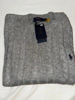 Grå kabelstickad tröja Polo Ralph Lauren - Snygg grå kabelstickad tröja från Polo Ralph Lauren i storlek M. Tröjan har rund hals, ribbade muddar och klassisk broderad logga på bröstet. Materialet är en mix av ull, akryl och nylon som gör den både mjuk och varm. Perfekt för kyliga dagar!
