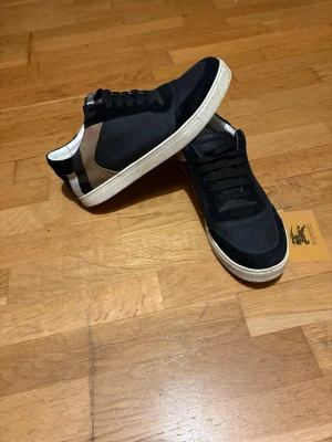 Burberry svarta sneakers med rutig detalj - Säljer ett par snygga Burberry sneakers i svart med klassisk rutig panel på sidan och vit sula. Skorna har snörning och rund tå, insidan är vit med Burberry-logga. Materialet är en mix av skinn och textil, perfekt för dig som gillar stilrena och trendiga sneakers.