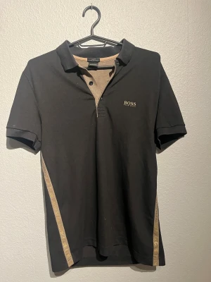 Svart polo från Hugo Boss - Säljer en svart polotopp från Hugo Boss med beige detaljer och logga på bröstet. Slim fit-modell med korta ärmar och klassisk krage. Materialet är bomull och har fuktavledande egenskaper. Snyggt inslag med beige band längs sidorna och knappslå framtill. 