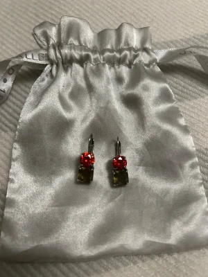 Dyrberg Kern siverplateterad  örhängen med  röda stenar  - Bilderna visar ett par örhängen med röda och olivgröna stenar, placerade på en silvrig satinpåse. 