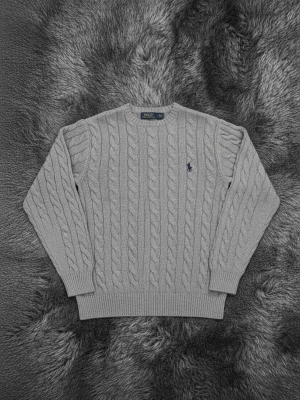 Grå kabelstickad tröja från Polo Ralph Lauren - Snygg grå kabelstickad tröja från Polo Ralph Lauren med klassisk rund hals och långärmad design. Tröjan har ett broderat marinblått logotyp på bröstet och ribbade muddar vid ärmslut och nederkant. Perfekt för dig som gillar stilrena och tidlösa plagg.