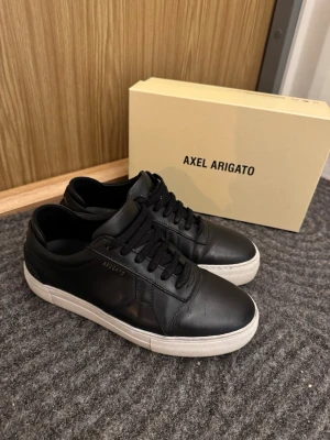 Svarta sneakers från Axel Arigato - Snygga svarta sneakers från Axel Arigato med vit platt sula och klassisk snörning. Skorna är tillverkade i skinn och har en stilren design med rund tå. Perfekta för dig som gillar minimalistisk och modern stil.