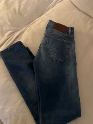 Blå jeans från G-Star RAW - Snygga blå jeans från G-Star RAW med klassisk femficksdesign och brunt läderpatch bak i midjan. Passar perfekt till sneakers och hoodie. 29-32