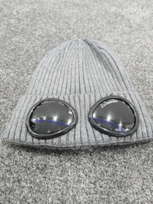 Grå mössa med glasögon från C.P. Company - Säljer en ribbstickad grå mössa från C.P. Company med två svarta glasögon-linser framtill. Mössan är tillverkad i mjuk ull och har en cool, urban vibe. Perfekt för dig som vill sticka ut med en unik accessoar.