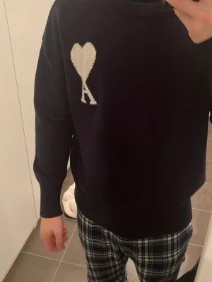 Mörkblå stickad tröja från Ami - Snygg mörkblå stickad tröja från Ami med vit hjärtapplikation och bokstaven A på bröstet. Tröjan har rund hals, ribbade muddar och är långärmad. Perfekt för dig som gillar stilrena detaljer och vill ha något som sticker ut lite.