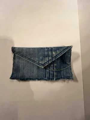 Blå denim plånbok med knapp - Säljer en liten kuvertväska i blå denim med råa kanter och metallknapp som stängning. Väskan har en enkel, rektangulär form och är perfekt för småsaker. Snygg detalj med synliga sömmar och klassisk jeanslook.