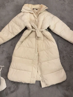 Lång beige/vit pufferjacka med bälte - Säljer en lång och mysig beige pufferjacka med bälte i midjan. Jackan har quiltad design, hög krage och dragkedja framtill. Perfekt för kalla dagar när du vill hålla dig varm och samtidigt se snygg ut. Kan eventuellt tänka mig gå ner i pris