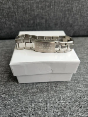 Maison Margiela silverfärgad armbandslänk - Säljer en chunky armbandslänk från Maison Margiela i silverfärgat rostfritt stål. Länken har ett coolt graverat nummermönster på spännet och breda, rektangulära länkar som ger ett stilrent och edgy uttryck. Perfekt för dig som gillar statement-accessoarer.