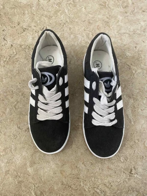 Adidas Campus sneakers svart/vit - Svarta Adidas Campus sneakers med vita ränder och vit sula. Klassisk låg modell med rund tå, platt sula och snörning. Ovandel i mocka och detaljer i syntet. Adidas-logga på plösen och CAMPUS-tryck på sidan. Perfekt för dig som gillar retrostil.