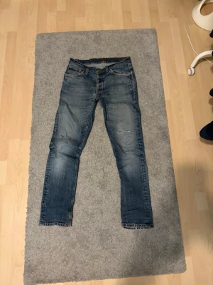 Blå jeans från Nudie Jeans, W31 L30 - Säljer ett par blå jeans från Nudie Jeans med klassisk femficksdesign och snygga slitningar. Jeansen har orangea sömmar och broderade detaljer på bakfickorna. Modellen är rak och passar dig som gillar en tidlös look.