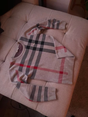 Rutig tröja från Burberry London - Säljer en snygg rutig tröja från Burberry London i grå med inslag av svart, grått och rött. Tröjan har rund hals, långa ärmar och klassiskt Burberry-mönster. Materialet är mjukt och stickat, perfekt för dig som gillar stilrena och ikoniska plagg.