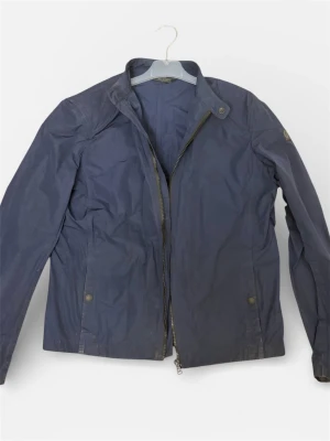 Blå vindjacka från Belstaff - Snygg marinblå vindjacka från Belstaff med klassisk ståkrage och dragkedja framtill. Jackan har långa ärmar, två fickor med tryckknappar och coolt Belstaff-märke på ärmen och insidan. Storlek är 16yr men passar S. Därav billiga priset. Jackan knappt använd och passar perfekt med hoodie under. PRIS kan diskuteras vid snabaffär