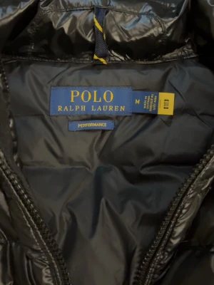 Svart dunväst från Polo Ralph Lauren - Säljer en svart dunväst från Polo Ralph Lauren med quiltad design och hög krage. Västen har dragkedja framtill och två sidofickor med dragkedja. Den är packbar och har en stilren, glansig finish som ger en modern look. Hör av dig vid funderingar. Använd fåtal gånger 🤗🤗