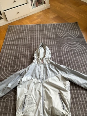 Nike vindjacka i ljusgrått, storlek L - Nike vindjacka i ljusgrå och vit färg med huva och dragkedja framtill. Jackan har två fickor med dragkedja och meshfoder på insidan för extra komfort. Perfekt för blåsiga dagar och sportiga outfits.