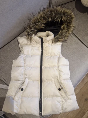 Vit dunväst - Säljer en vit dunväst från H&M med huva och fuskpälsdetalj. Västen är otroooligt vacker blir ledsen att behöva sälja den, har quiltad design, två fickor med knappstängning och dragkedja framtill. Perfekt för kyliga dagar när du vill vara både varm och snygg.