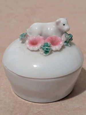 Liten rund förvaring med lock - Bilderna visar en liten rund förvaring med lock, dekorerad med en vit figur av ett lamm, rosa blommor och lite grönt. Kan användas som dekoration och förvaring 