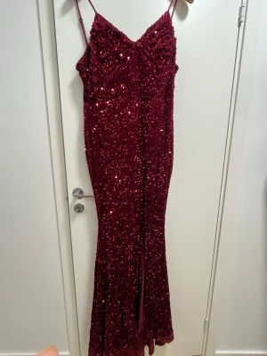 Vinröd glittrig långklänning - Säljer en vinröd långklänning med tunna axelband och glittriga paljetter över hela klänningen. Klänningen har en figurnära passform och en slits framtill som ger en snygg siluett. Perfekt för dig som vill sticka ut med en glamorös look.