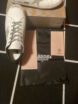 Golden Goose Superstar sneakers vit/grå - Golden Goose Superstar sneakers i vitt skinn med grå mockadetaljer och silverfärgad stjärna på sidan. Skorna har rund tå, platt sula och grå snörning. Märkets logga syns på hälen och innersulan. Snygg och stilren design med GGDB-detalj på sidan.