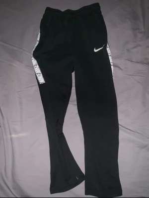 Svarta träningsbyxor från Nike - Svarta träningsbyxor från Nike med vita detaljer längs sidorna och en vit Nike-logga på vänster ben. Byxorna har elastisk midja och är tillverkade i ett mjukt syntetmaterial som passar perfekt för sport och träning. Storlek 147-158 så typ xxs/xs
