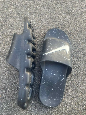 Nike svarta sandaler med stänkmönster - Unika svarta sandaler från Nike med vitt stänkmönster och stor Nike-logga på ovansidan. Sulan har en cool, upphöjd design som påminner om sneakers och ger extra komfort. Perfekt för chill dagar och enkel att slänga på sig. Materialet är syntet och färgen är svart med vita detaljer.