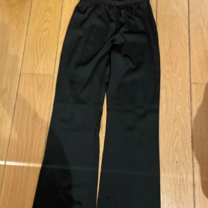 Svarta byxor från Ahlvar Gallery - Snygga svarta byxor från Ahlvar Gallery i storlek XL. Byxorna har en resår i midjan och rak snitt som ger en avslappnad och trendig look. Materialet är silkigt och har en lätt glans, perfekt för dig som gillar stilrena och bekväma plagg. Snygg både till fest och vardags! 