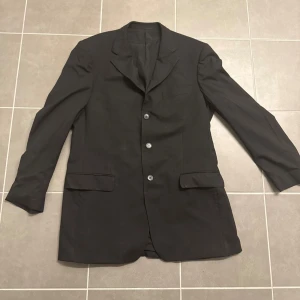 Svart kavaj från Ermenegildo Zegna - Stilren svart kavaj från Ermenegildo Zegna med klassisk krage och tre knappar framtill. Kavajen har två fickor med lock och en bröstficka. Materialet känns mjukt och har en elegant finish, perfekt för dig som vill ha en snygg och tidlös look. Storlek 52, Armlängd 67 och Axelbredd 51