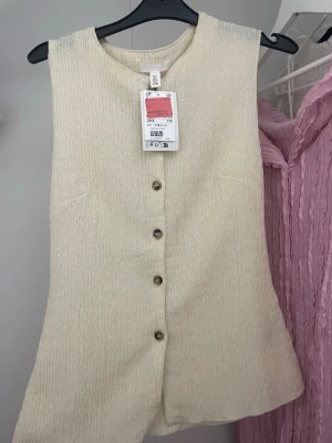 Beige ärmlös blus med knappar - Säljer en beige ärmlös blus från H&M med strukturerat tyg och rund sopringning. Blusen har dekorativa knappar framtill och en rak passform som ger en clean look. Perfekt att styla med jeans eller kjol för en enkel men snygg outfit.
