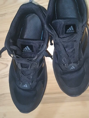 Svarta Adidas sneakers med snörning - Säljer ett par svarta Adidas sneakers med klassiska tre ränder på sidan och logga på plösen. Skorna har rund tå, platt sula och är gjorda i syntetmaterial och textil. Perfekta för dig som gillar sportig stil och vill ha bekväma skor med snygg design.