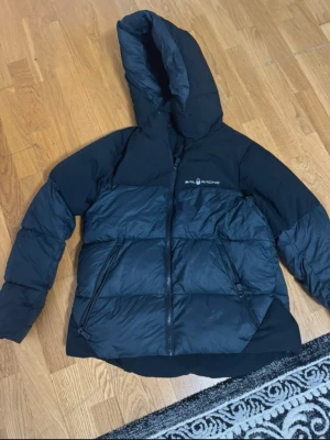 Svart pufferjacka från Sail Racing - Svart pufferjacka från Sail Racing med huva och två snedställda fickor med dragkedja. Jackan har en stilren look och är perfekt för kalla dagar. Materialet är syntet och jackan har en quiltad design som ger extra värme. Snygg logga på bröstet.