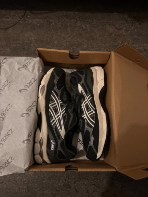 Svarta Asics Gel sneakers med snörning - Säljer ett par svarta Asics Gel sneakers med vita och grå detaljer. Skorna har en chunky sula, mesh och syntetmaterial i ovandelen samt klassisk snörning. Ikoniska Asics-loggan på sidan och GEL-text bak på hälen. Perfekt för dig som gillar sportig stil. Helt nya! Och storlek 42 för dig som har små ben och inte så breda så är dom här skorna perfekta för dig!