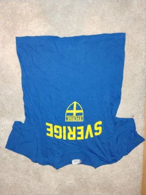 Blå Sverige t-shirt AB Karl Hedin - Blå t-shirt med tryck i gult där det står 'Sverige' och en svensk flagga på framsidan samt 'AB Karl Hedin' med trädgrafik på ryggen. Klassisk rund hals och korta ärmar, perfekt för dig som vill visa lite svensk stolthet.
