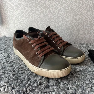 Bruna Lanvin sneakers i mocka - Lanvin sneakers i brun mocka med grön tå. Snygga bruna snören och vit platt sula med Lanvin-logga undertill. Kommer med original dustbags. Säljer då dom blivit för små. Skorna är använda men i gott skick. Enda defekten är att dom är slitna inuti hälen som man kan se på bilderna. 