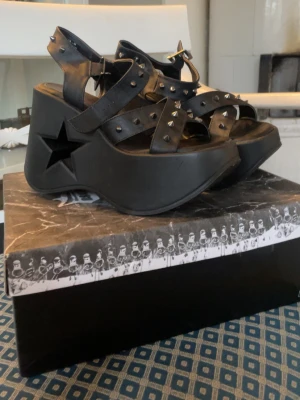 Svarta sandaler med nitar och platå - Säljer ett par svarta sandaler från Demonia i veganskt skinn med coola silverfärgade nitar och kraftig platåsula. Sidan på sulan har en utskuren stjärna och remmarna har spänne. Perfekt för dig som gillar edgy stil och vill sticka ut.