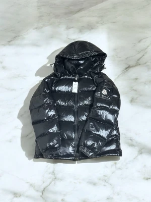 Svart pufferjacka från Moncler äkta  - Svart pufferjacka från Moncler med glansig finish och huva. Jackan har dragkedja framtill och Moncler-logga på ärmen. Fylld med dun och har en quiltad design som ger extra volym och värme. Perfekt för kalla dagar och riktigt snygg streetstil.
