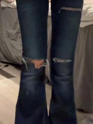 Bootcut jeans med slitningar, mörkblå - Snygga bootcut jeans i mörkblå tvätt med slitna detaljer vid knäna och låren. Jeansen har breda ben och klassisk femficksdesign. Materialet är jeans och de har en cool, lite vintage vibe med de slitna partierna framtill. Jeans ifrån gina 14+ nästan aldrig använda ena hållet har töjts ut lite som man ser på bilden men annars jättefint skick ❣️storlek S